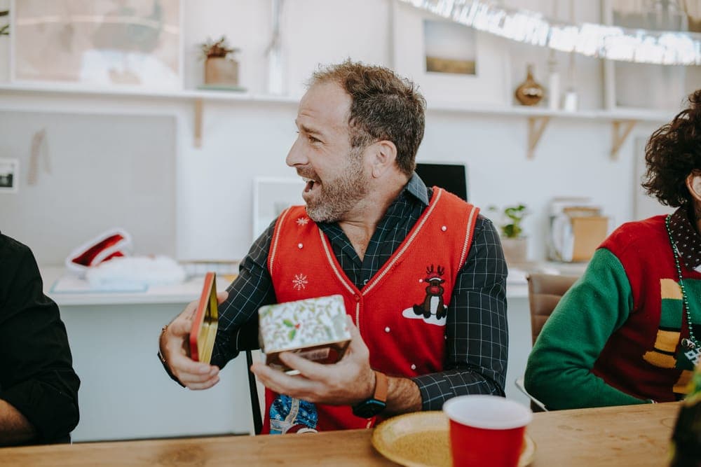 A man in a holiday sweater opening a gift at home