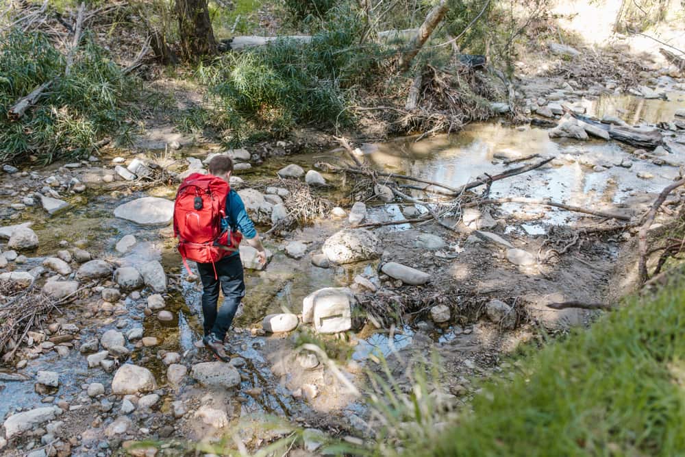 A hiker with a red backpack crossing a rocky stream