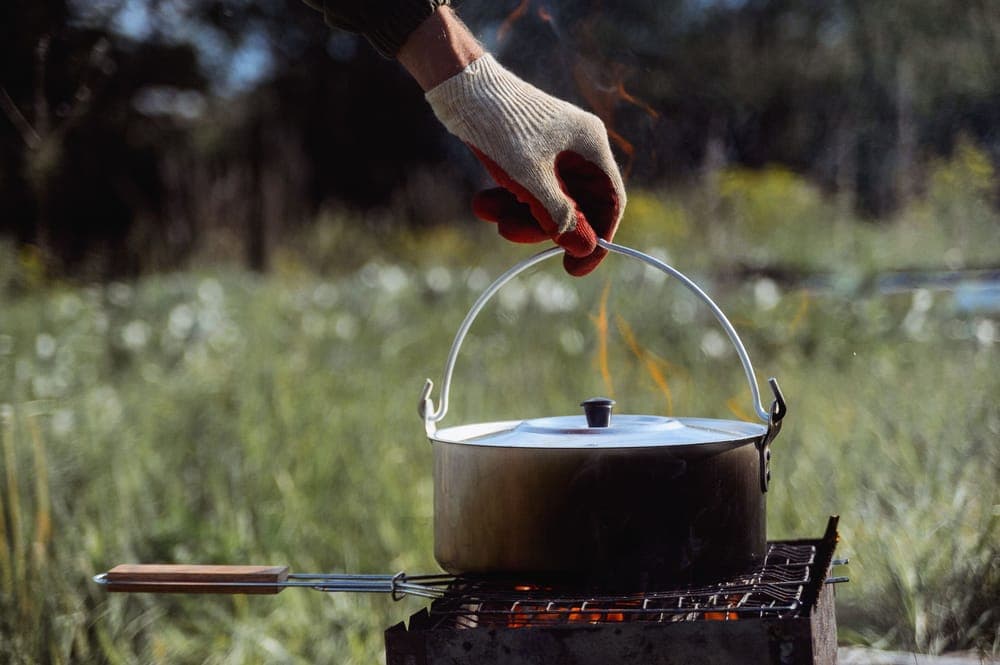 Illustrative stock photo: a gloved hand holding a pot over a campfire grill