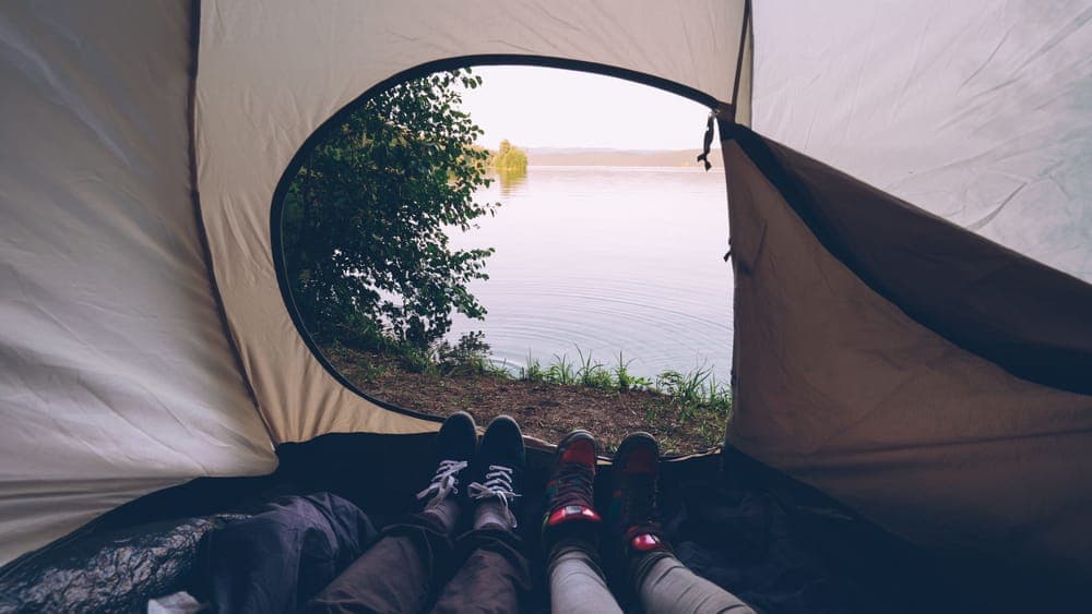 Illustrative stock photo: the view from a tent toward a calm lake with two people nearby